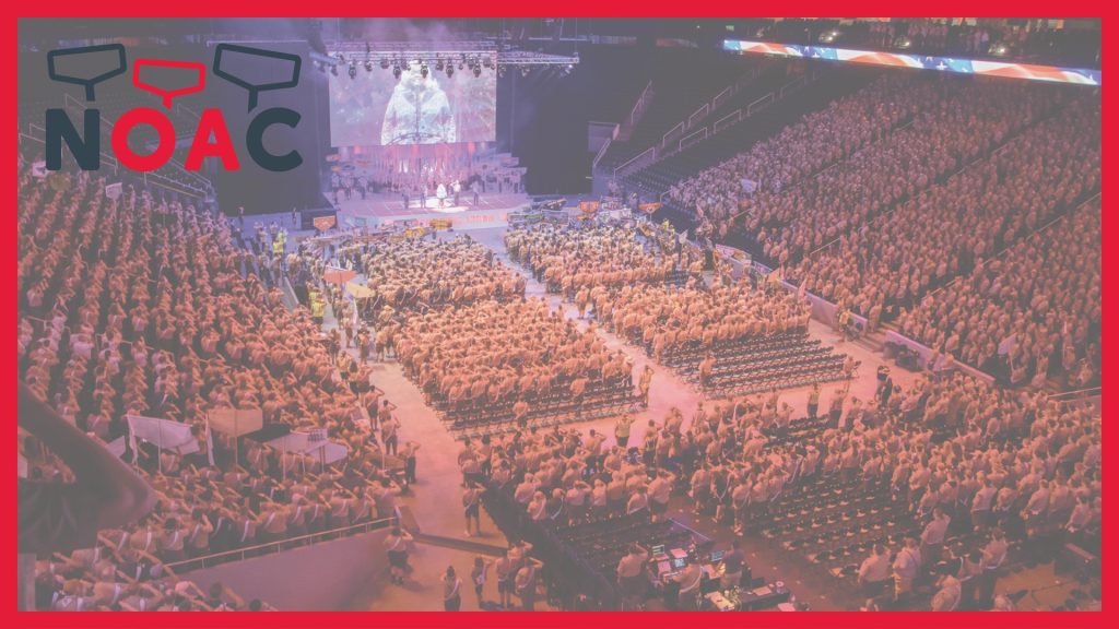 A large crowd of people in a conference arena with a stage and screen displaying a speaker. The crowd is wearing matching uniforms and some are holding flags. The words "NOAC" are displayed at the top.