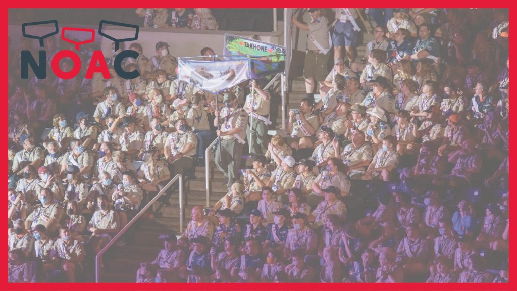 A crowded indoor event with rows of people in scout uniforms sitting on tiered seats, some holding banners, with ambient stage lighting, and a large 'NOAC' logo at the top.