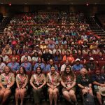 A large, diverse group of people, many wearing colorful shirts and uniforms, sit in an auditorium with tiered seating. The front row features individuals in scouting uniforms, smiling at the camera. The well-lit theater is filled with rows of attendees.